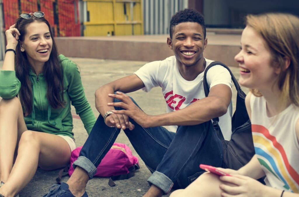Three older teens, aged 17-18, sitting on ground with backpacks, talking and laughing.