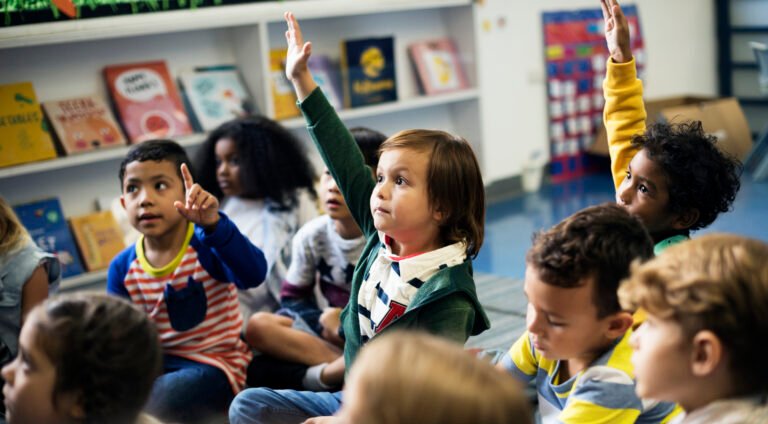 Group of very young students, aged 5-6, sitting on carpet of school library all facing the same direction, as if listening to someone speak.  Two students raise their hands high.  Books displayed on shelves in the background.
